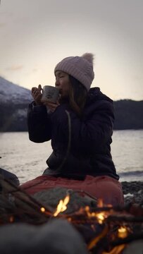 Woman sitting near campfire enjoying cup of hot drink