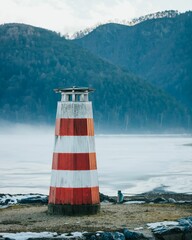 Vertical shot of a red and white lighthouse on the shore against the background of green hills. - obrazy, fototapety, plakaty