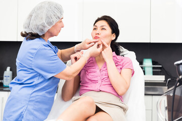Experienced woman cosmetologist examining lips of asian female client in aesthetic medicine clinic...