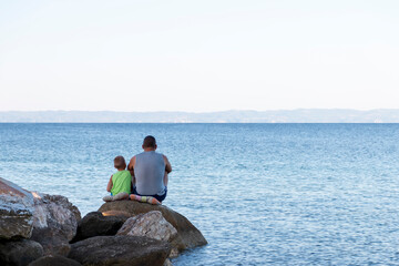 Father and child sitting on the sea coast  and enjoying sunny beautiful morning travelling together. Father and lovely son  resting in the sunrise