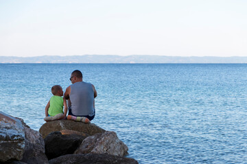 Fototapeta na wymiar Father and child sitting on the sea coast and enjoying sunny beautiful morning travelling together. Father and lovely son resting in the sunrise