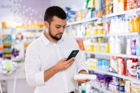 Portrait of focused bearded man choosing cosmetic product at pharmacy, using smartphone for scanning barcode