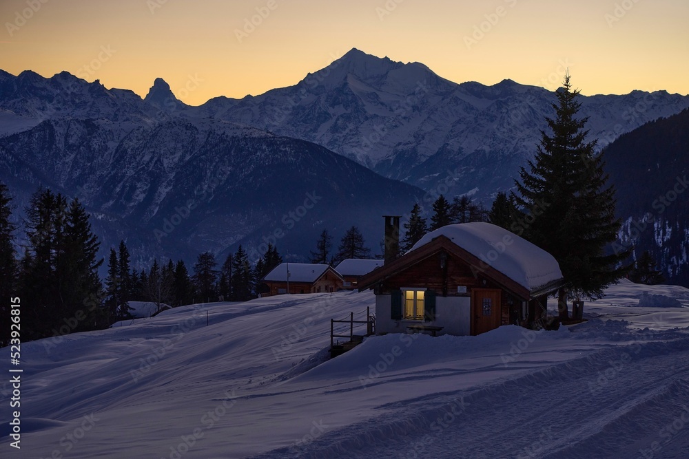 Sticker Scenic view of a wooden cottage in a snowy mountain at night with the orange sky in the background