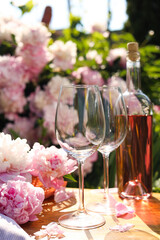 Bottle of rose wine and glasses with beautiful peonies on wooden table in garden