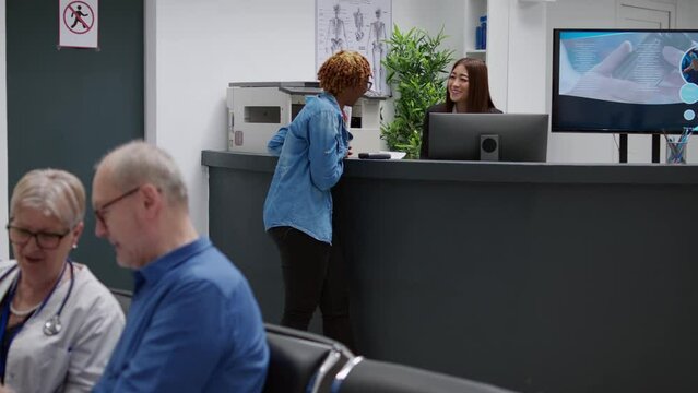 Patient talking to receptionist at hospital reception desk, asking for help with medical appointment and treatment. African american woman chatting with clinical staff in waiting area lobby.