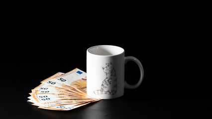 Banknotes of fifty euros, arranged in a fan, under a white cup decorated with a cat, all on a black background