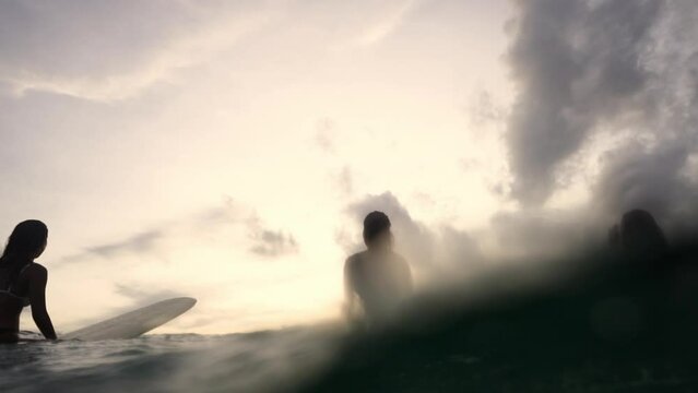 Low angle view of Asian woman surfer paddling surfboard and riding the wave in the sea at tropical beach at sunset. Girl friends enjoy outdoor lifestyle and water sport surfing on summer vacation