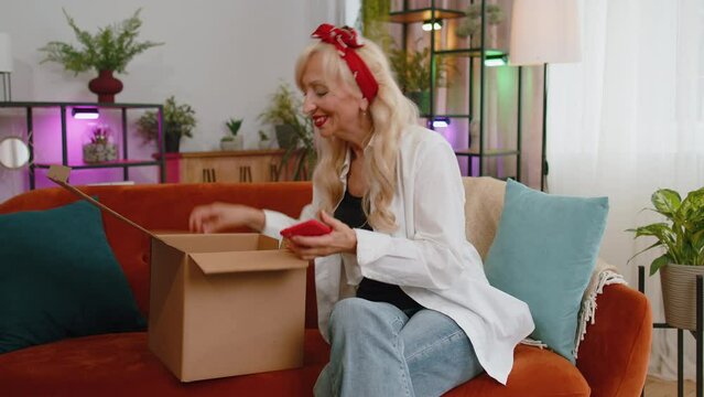 Happy senior grandmother unpacking delivery parcel sitting at home. Smiling satisfied elderly woman shopper, online shop customer opening cardboard box receiving purchase gift by fast postal shipping