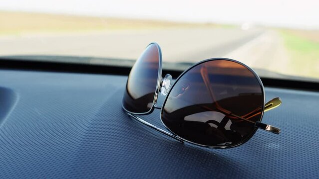 Modern folded sunglasses lie on car dashboard panel against moving wipers macro. Automobile stands on side of asphalt road on sunny day