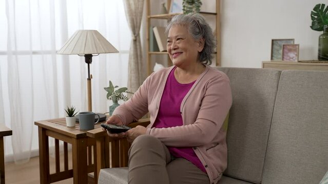 leisure asian retired senior woman enjoying watching a funny show after tuning to the channel with tv controller in the living room at home