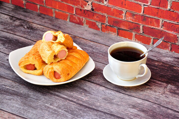 Grilled sausages in puff pastry and a cup of black coffee on a wooden table against a brick wall.