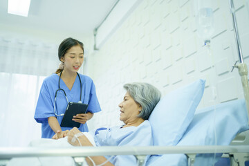 Senior Asian female patient resting on the medical bed in hospital and talking to doctor. 