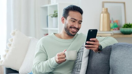 Browsing on his phone, relaxing on the sofa and drinking coffee while sitting at home on the weekend. Young man feeling happy, positive and smiling while reading a text message or seeing a funny meme