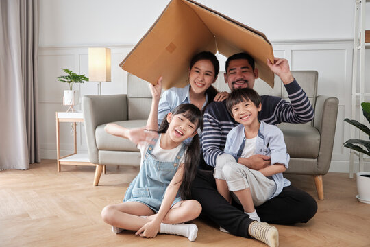 Asian Thai family, adult dad, mum, and little children happily fun playing in a white living room, built cardboard paper safety house with imagination, lovely weekend and wellbeing domestic lifestyle.