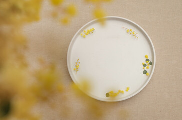 Warm background texture of beige table with a white round plate or tray. Wedding stationery mockup scene. Plant branches with beautiful golden wattle flowers. Copy space for text, card, or product.