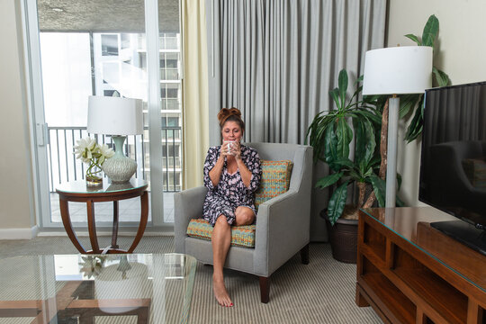 Beautiful Middle Aged 50-year-old Woman In A Robe Relaxing And Enjoying A Cup Of Morning Coffee Or Tea On A Comfortable Chair In A High Rise Apartment Home