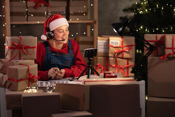 Young attractive caucasian woman dressed as santa claus takes an order online using headset and...