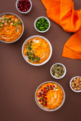 Pumpkin puree soup of butternut squash in bowls with chickpeas and green onions, pumpkin and pomegranate seeds, top view on plates with food on brown background