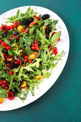 Summer delicious salad with arugula and fresh berries, red and black currants, sweet cherries and black mulberries, salad in an oval white plate on a green background