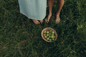 legs of a young woman in a long skirt and legs of a man on green grass. the couple were picking apples in the garden and put them in a wicker bowl. bowl with green apples on the grass