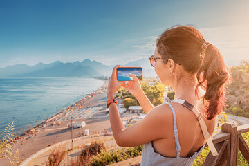 Fototapeta premium Travel blogger girl takes high-quality and vivid photos on the camera of his new expensive smartphone of the famous Konyaalti beach from a scenic viewpoint in Antalya, Turkey