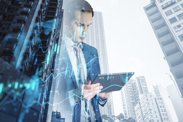 Attractive young european businessman using tablet with creative growing financial forex chart graph and map on blurry city background. Market, stock and trading concept. Double exposure.