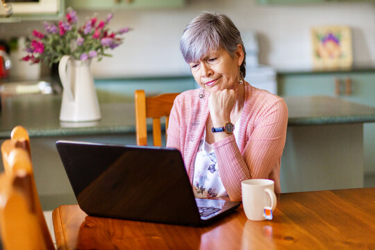 Senior woman with cup of tea working on laptop from home