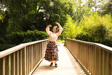 Beautiful teenage flamenco dancer dressed in a flowery skirt with ruffles and flowers in her hair is dancing in the park on a wooden bridge. Flamenco concept cultural heritage of humanity.