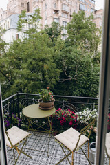 a coffee table and two chairs on an open balcony with a city view. blooming flowers in pots decorate the balcony