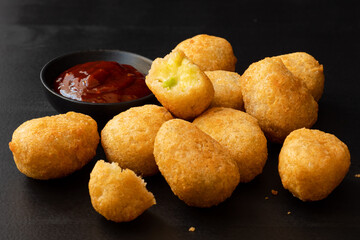 Fried breaded chilli cheese nuggets next to a bowl of ketchup on dark background. One eaten.