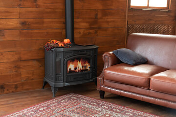 Black modern Cast iron wood stove at home. Fragment of interior of country house. Interior cozy living room with wood burning stove inside, comfortable sofa, rustic wooden walls and carpet on floor.	