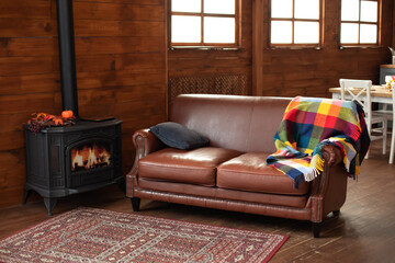 Interior cozy living room with wood burning stove inside, comfortable sofa, rustic wooden walls and carpet on floor. Black modern Cast iron wood stove at home. Fragment of interior of country house. 
