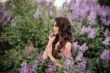 Fototapeta premium a beautiful girl with makeup and hair styling in underwear stands in a garden with lilacs on a summer evening