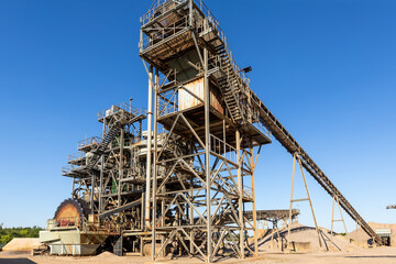 Fototapeta na wymiar Panoramic view heavy metal construction open pit gravel plant sand quarry big rusty rotor machine material excavating and crushing on blue sky. Industrial machinery factory equipment steel background