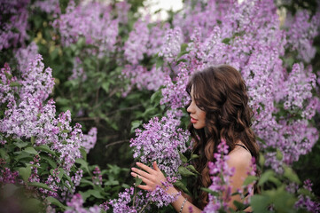 Fototapeta premium a beautiful girl with makeup and hair styling in underwear stands in a garden with lilacs on a summer evening