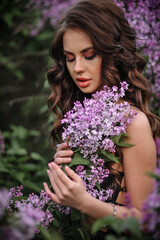 a beautiful girl with makeup and hair styling in underwear stands in a garden with lilacs on a summer evening