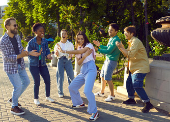 Bunch of happy multiracial friends meet up at the city park in summer. Multi ethnic group of cheerful, joyful, excited young people dancing, having fun and enjoying good time outdoors together