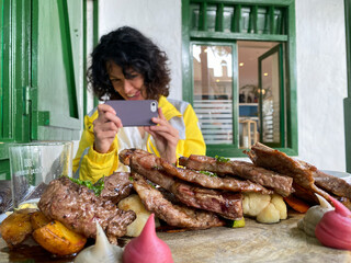 Smiling young female food blogger clicking photo of steak on restaurant counter using smart phone