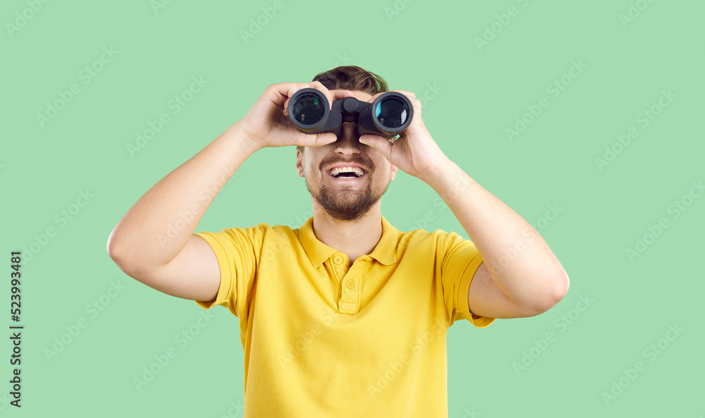 Wall mural Happy joyful young man in casual yellow T shirt standing isolated on green background, holding modern binoculars, looking ahead and smiling. Making discoveries and having fun while travelling concept