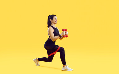 Fototapeta na wymiar Smiling young toned female fitness instructor do squats with dumbbells and bands on yellow studio background. Motivated happy fit woman athlete workout train alone. Sport lifestyle.