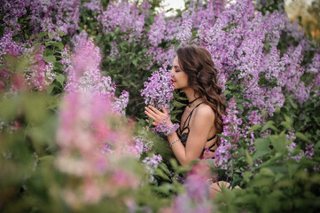 a beautiful girl with makeup and hair styling in underwear stands in a garden with lilacs on a summer evening