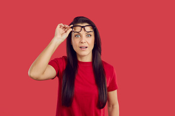 Portrait of stunned young woman isolated on red studio background take off glasses shocked by amazing sale offer or deal. Amazed female surprised with unbelievable news or discount. Wow face.