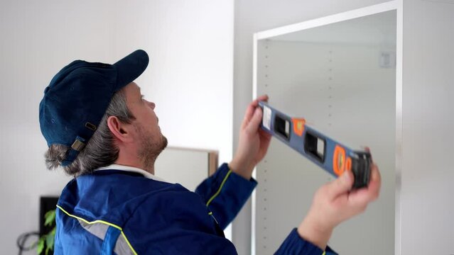 Worker installs cabinet frame in room. Man in uniform measure the wardrobe and rejoices successful assembly