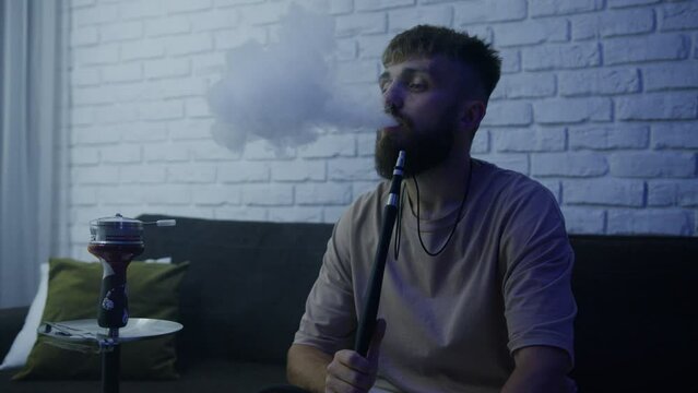 Smoking hookah at home. A man exhales smoke while sitting on the couch