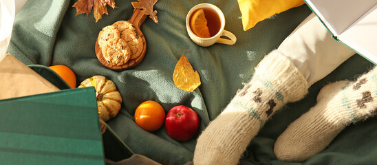Woman in knitted socks, with book sitting on plaid with food and cup of tea, top view