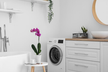 Stylish bathroom interior with washing machine and orchid flower on table near white wall