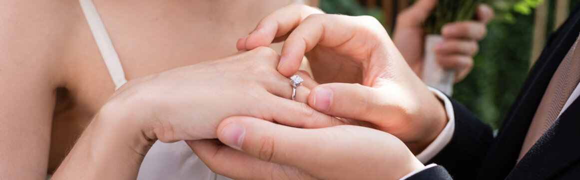 Cropped view of groom wearing ring on finger of bride outdoors, banner.