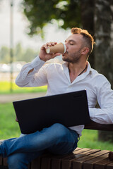 Happy man works in the park with a laptop, drinks coffee. A young man on a background of green trees, a hot sunny summer day. Warm soft light, close-up.