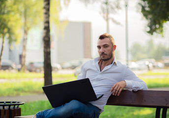 A handsome business man in a white shirt is working in the park with a laptop. A young man on a background of green trees, a hot sunny summer day. Warm soft light, close-up.