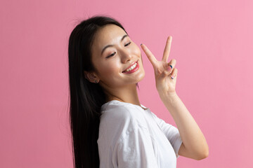 Portrait of asian positive smiling woman with V fingers gesture on pink background.